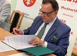 Marszałek Adam Struzik w okularach, siedzi za stołem prezydialnym, pochylony nad teczką z dokumentami w pozie składającego podpis.