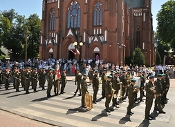Żołnierze z orkiestry wojskowej stoją na pustej ulicy przed katedrą. Ustawieni są w rzędy