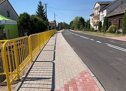 Prosty odcinek pustej drogi w szerokim planie, ujęcie z prawej strony z chodnika z żółtą barierką po lewej.
