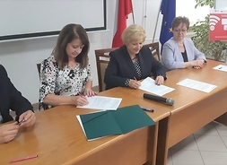 Cztery osoby za stołem prezydialmym, siedzą i podpisują dokumenty.