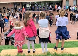 Czworo dzieci szkolnych stoi tyłem w rzędzie, za nimi dziedziniec przed budynkiem pełen uczestników uroczystości.