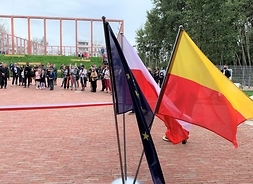 Trzy flagi: Polski, Unii Europejskiej i m.st. Warszawy powiewają na dzidzińcu, za nimi w oddaleniu grupa uczestników wydarzenia.