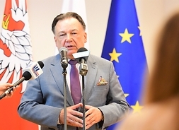 Marszałek stoi przed mikrofonem i mówi, za nim flagi Polski, UE i Mazowsza.