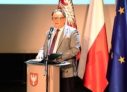 Marszałek stoi na trybunie, mowi do mikrofonu, za nim flagi Polski, UE i Mazowsza.