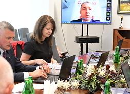Wicemarszałek Rajkowski i członek zarządu Janina Ewa Orzełowska siedzą obok siebie przy stole. Oboje parzą w monitory otwartych laptopów