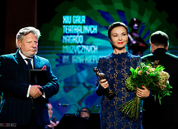 Mężczyzna w garniturze galowym stoi na scenie obok kobiety w sukni wieczorowej. Kobieta trzyma w prawym ręku statuetkę, w lewym bukiet kwiatów. Oboje patrzą się przed siebie i uśmiechają się