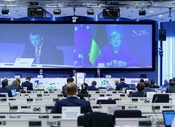 w ogólnym planie sala obrad Komitetu Regionów w Brukseli, uczestnicy patrza na wielki ekran na którym z lewej prowadzący obrady, z prawej kanclerz Angela Merkel..