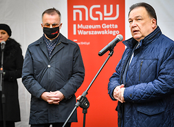Mrszałek Adam Struzik mówi do mikrofonu stojąc, za nim banner z logotypem Muzeum Getta Warszawskiego. Po lewej stoi męzczyzna w pozie skupienia, obok stoi kobieta.