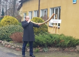 Wój gminy Wiesław Michalczuk stoi na zewnątrz przed budynkiem szkoły. W rękach trzyma długi róg, który przytyka do ust.