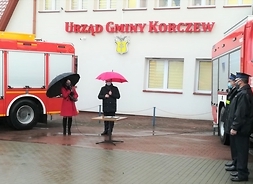 Na tle ściany budynku urzędu gminy: dwa pojazdy strażackie, pomiędzy stoją kobieta i mężczyzna pod parasolami.