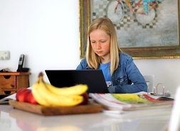Dziewczynka siedząca przed otwartym laptopem, wpatrzona w ekran. Przed nią na stole stoi talerz z bananami i innymi owocami.