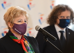 stoją obok siebie dwie osoby, z lewej strony na stojąco do mikrofonu przemawia członek zarządu województwa mazowieckiego Elżbieta Lanc w maseczce