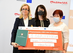 Członek zarządu Janina Ewa Orzełowska trzyma wielki symboliczny czek. Po jej prawej i lewej stronie stoją dwie przedstawicielki urzędu gminy w Michałowicach. Także trzymają czek