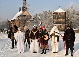Grupa kolędników w przebraniach na tle zabudowań skansenu w Sierpcu. Zdjęcie w zimowej scenerii.