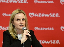 kobieta w pozycji siędzącej  mikrofonem w ręku, w tle banser w kolorze czerwonym z białym napisem Mazowsze serce Polski