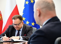 Marszałek, siedząc przy stole, podpisuje dokumenty. Za nim są flagi Mazowsza, Polski i Unii Europejskiej