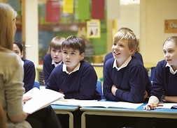 Trójka uczniów słucha nauczycielki, siedząc w szkolnych ławkach