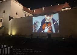 Kadr z prezentacji multimedialnej wyświetlanej na murze budynku przedstawiający starszego mężczyznę grającego na skrzypcach.