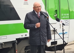 Waldemar Kuliński stoi przed mikrofonem i przemawia. Za nim znajduje się pociąg kolei w barwach kolei mazowieckich