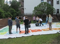Grupa uczniów na dużej planszy rozłożonej na trawie. Jeden toczy olbrzymią kostkę do gry