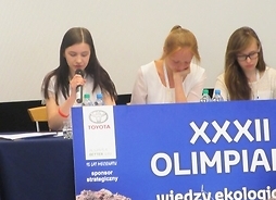 Trzy finalistki przy stole z tabliczką XXXII Olimpiada, jedna z mikrofonem w ręku