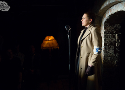 scena z przedstawienia - aktorka stoi przy mikrofonie w jasnym płaszczu i opaską z gwiazdą Dawida na ramieniu