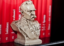 Niewielkie gipsowe popiersie marszałka Józefa Piłsudskiego na tle książek w czerwonych okładkach.