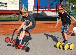 Dwóch chłopców około 10-letnich jadących na wózkach z kółkami, rodzaju hulajnogi, ubranych w pióropusze
