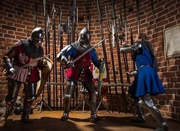 Sala w zamku, troje inscenizatorów przebranych za rycerzy, w tle halabardy