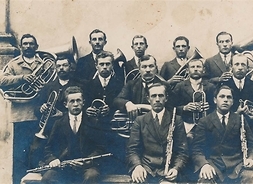 Grupa muzyków siedząca w dwóch rzędach w odświętnych ubraniach