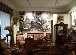 Fragment wystawy prezentujący salon Prezydenta - krzesła i stół. Nad nimi zdjęcie historyczne Mościckiego z gośćmi