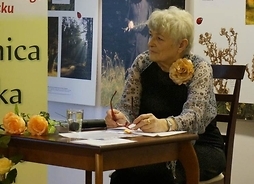 Pisarka siedzi przy małym stoliku w bibliotece. Na stole kartki i dwie róże