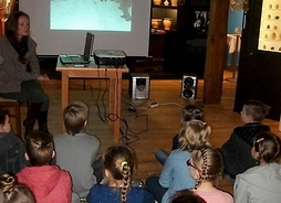 Grupa dzieci siedzi w pomieszczeniu muzealnym i ogląda projekcję filmu z komentarzem siedzącej przy rzutniku prowadzącej