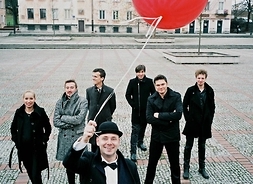 Zespół w strojach jesiennych na wyłożonym kostką brukowa placu miejskim. Jeden z muzyków, stojący na pierwszym planie, trzyma w ręku czerwony balonik