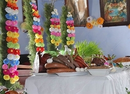 Stół zastawiony potrawami świątecznymi – kiełbasami, jajami, wędliną, udekorowany pisankami. W tle ozdoby Wielkanocne z papierowych kwiatów