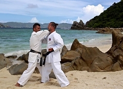 Dwóch karateków ćwiczących układ w kimonach na plaży. W tle morze i widoczny półwysep wyspy Okinawa