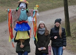 Trzy dziewczyny na wiejskiej drodze, jedna niesie dużą kukłę Marzanny na kiju, wykonaną z materiałów i krepin