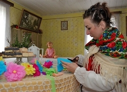 Artystka w stroju ludowym w barwnej chuście wycina papierowe kwiaty siedząc przy stole we wnętrzu zabytkowej chałupy