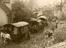 Stara fotografia przedstawiająca ciąg romskich wozów konnych przejeżdżających przez błotnistą drogę na obrzeżach miasta