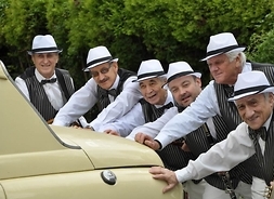 Zespół w białych koszulach, kamizelkach i kapeluszach pchający samochód z lat 60. XX w.