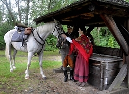 Aktor w stroju ułana, aktorka w stroju ludowym i koń z rzędem przy zabytkowej, drewnianej studni