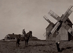 Chłop ubrany w mundur, obok żona na koniu, w tle dwa drewniane wiatraki i gospodarstwo rolne