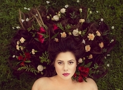 Portret piosenkarki leżącej na trawie z licznymi kwiatami wplecionymi w rozpuszczone, sztucznie przedłużone włosy
