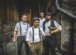 Czterech muzyków w ubraniach stylizowanych na przedwojennych muzykantów (szelki, kaszkiety, koszule z podwiniętymi rękawami i kamizelki) na obdrapanym podwórku kamienicy