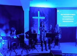 Trzy panie z mikrofonami i dwóch gitarzystów na scenie, w tle duży krzyż i wyświetlacz z tekstem religijnej piosenki