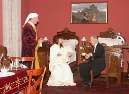 Wnętrze secesyjnego pokoju – stół z zastawą, krzesła, łóżko, komody – na krzesłach aktorzy przebrani w stroje z epoki