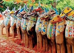 Kilkanaście glinianych figur koni z rzędem, pomalowanych w geometryczne wzory stojących na ziemi zasypanej płatkami kwiatów