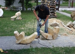 Rzeźbiarz dużą wiertarką przygotowuje otwory we fragmencie rzeźby. Obok leży drugi fragment, w tle kolejne części abstrakcyjnej konstrukcji