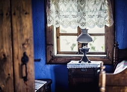 Wnętrze chałupy, widoczne głównie drewniane okno ze zdobioną firanką i lampa naftowa na stoliku