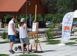 Dwoje młodych ludzi w letnich strojach z wózkiem dziecięcym stoją przed sztalugami na których jest kartka z informacją o wydarzeniu. W tle powiewająca flaga z napisem Dni Otwarte Funduszy Europejskich
